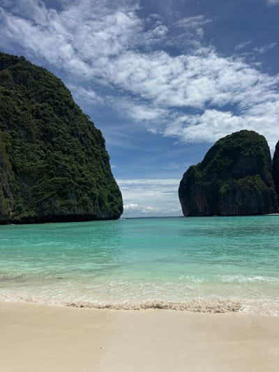 This image depicts a scenic view of Maya Bay in Ao Nang, Krabi Province, Thailand. The foreground features a pristine white sand beach meeting calm, turquoise waters. Gentle waves lap at the shore, creating a foamy white line. In the midground, the expansive turquoise sea stretches towards the horizon, dotted with two towering limestone karsts covered in lush green vegetation. These karsts rise dramatically from the water, framing a gap that leads to a distant view of more islands. The sky above is a vibrant blue, adorned with scattered white, fluffy clouds. The overall impression is one of a serene and breathtaking tropical paradise.