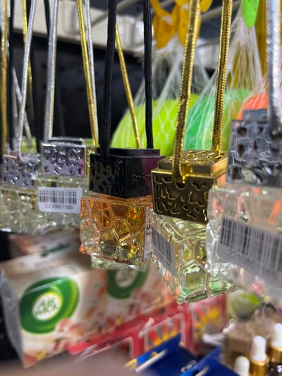 The image depicts a close-up view of multiple car air fresheners or diffusers suspended for retail display in a commercial establishment in Alasia, Nigeria. The primary subjects are small, glass bottles, each containing a translucent liquid and topped with a decorative cap and a hanging string.
Specifically, there are at least eight individual units visible, arranged in rows. Each unit consists of a clear glass bottle, approximately 5-7 cm tall, filled with either clear or amber-colored liquid. Some bottles feature a distinctive hexagonal, honeycomb-like texture, while others appear smoother or have ridged sides. Attached to the top of each bottle is a metal or plastic cap, presented in silver, gold, or black finishes. These caps often display intricate cut-out patterns or embossed designs. A thin, braided string, matching the cap's color (silver, gold, or black), extends upwards from each cap for suspension.
Several bottles bear white rectangular price or inventory stickers with black barcodes and alphanumeric codes, such as "2210801165." One black-capped unit clearly displays the text "Calvfor" (likely "Calvin") on its side, suggesting a brand identifier.
The items are suspended from an unseen structure, likely a display rack or shelving unit, against a blurred background indicative of an indoor retail environment. Visible in the soft-focus background are other consumer products, including a white package with a prominent green "Air Wick" logo, further confirming a store setting. Brightly colored objects in green, yellow, and orange, possibly other products or decorations, are also present in the background. No individuals are visible, and no actions or direct interactions are occurring within the frame; the scene is static. The lighting is consistent with interior artificial illumination.