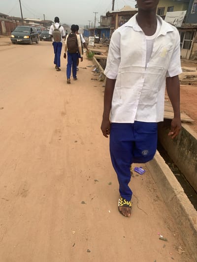 The image depicts a street scene in Shagamu, Nigeria. In the foreground, a person wearing a white short-sleeved shirt over a grey t-shirt and blue trousers walks away from the camera. They are wearing yellow and black slide sandals. To the left of the foreground subject, three other individuals, likely students due to their matching uniforms, are also walking away from the camera. They are wearing white shirts, blue trousers, and backpacks. The individuals are walking on a dirt road, with a concrete drainage ditch to the right. Several vehicles, including a dark-colored minivan and other cars, are visible further down the road. The background consists of buildings and structures that suggest a bustling urban environment. The overall lighting suggests an overcast day.