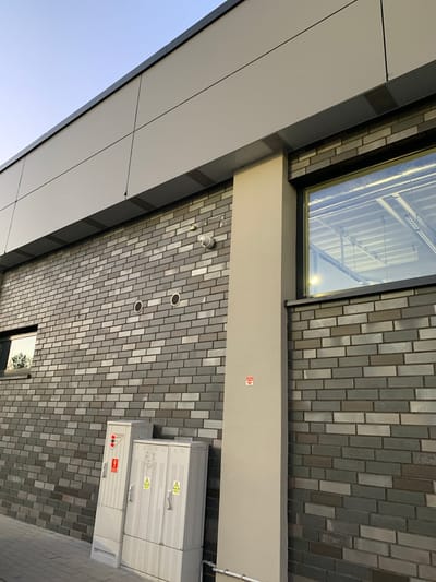 The media file is a low-angle shot of a modern building facade, likely in Białystok, Poland. The building features a combination of materials: a light-colored, panelized section on the left contrasts with a dark-colored section and a wall clad in grey brick tiles. Above, a large window allows a view into an interior space with overhead lighting and structural elements.
Mounted on the brick facade are several objects. A white security camera is visible near the top left of the tiled section. Further down, two circular ventilation grates are set into the brick. On the right side of the image, two grey electrical cabinets are mounted on the wall. The upper cabinet has two yellow warning signs with black exclamation marks and is divided into two doors. The lower cabinet is a longer, single-door unit with a red and white label displaying electrical symbols.
A small red and white sticker with an arrow pointing right is affixed to a light-colored horizontal beam that spans across the image, separating the upper window area from the brick facade.
The sky is a clear, pale blue, suggesting it is daytime, possibly late afternoon or early morning given the lighting. The overall scene appears clean and functional, with no people or animals visible. The building's design suggests a contemporary urban setting.