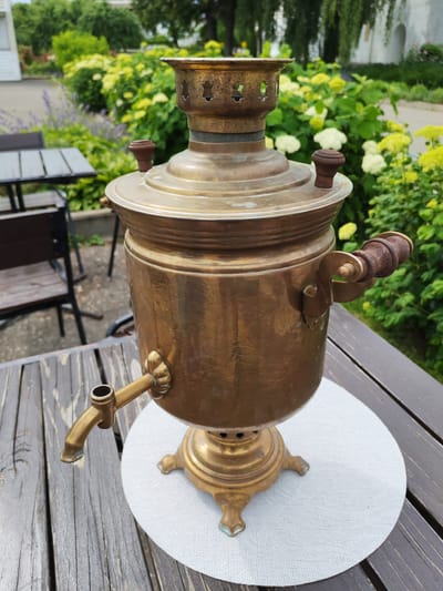 A brass samovar sits on a wooden table, outdoors. The samovar is ornate, with decorative handles and a spigot. It is placed on a white placemat. In the background, lush green bushes and flowers are visible, along with some outdoor seating areas and tables. The scene appears to be in a garden or courtyard setting, possibly a cafe or restaurant, given the presence of the tables and chairs. The lighting suggests it is daytime, with bright but not harsh sunlight, indicating pleasant weather. The overall impression is one of a serene, possibly historic, outdoor setting. There is no visible text in the image.