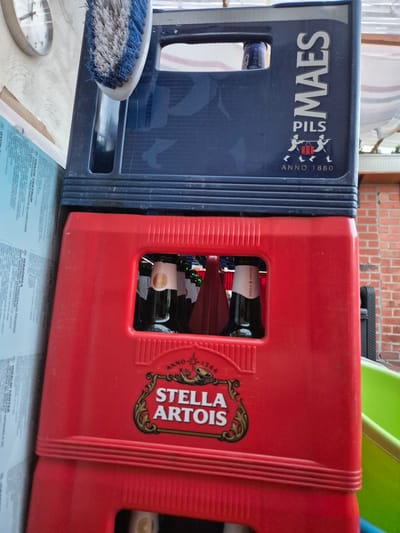 The image shows two beer crates stacked vertically. The top crate is dark blue and has "MAES PILS" printed on its side, along with "ANNO 1880". The bottom crate is red and displays the "Stella Artois" logo. Several bottles of Stella Artois beer are visible through the opening in the red crate. A blue and white brush is resting on top of the blue crate. In the background, a part of a wall clock is visible on the left, and a brick wall is seen on the right. The scene appears to be outdoors, possibly in a backyard or a storage area. There is no discernible location in the city of Aalst, Belgium.
