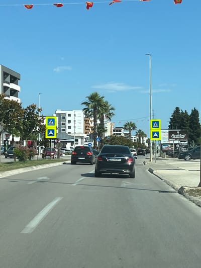 The image shows a street scene in Ulcinj, Montenegro, under a clear blue sky. Red and yellow flags are strung across the top of the image. On the left side of the road, there is a multi-story modern building with balconies and trees in front. On the right side of the road, there is a street light and several signs, including two yellow pedestrian crossing signs. Further down the road, there are palm trees, more buildings, and a gas station. Several cars are on the road, including a black Mercedes-Benz sedan in the foreground, a black Volkswagen Golf, and other vehicles further ahead. The road has white lane markings. The scene appears to be a typical urban environment with commercial activity.
