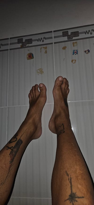 The image captures two bare human legs and feet, viewed from a top-down perspective, resting on a tiled floor. The person has dark-toned skin. Both legs display tattoos. On the upper right leg, there is a tattoo resembling a starburst or compass rose with a trailing dotted line. The lower left leg features a tattoo of a stylized bird or creature with an 'X' and what might be a heart. A partially visible tattoo on the upper left leg suggests text, possibly "Cob".
The setting is an indoor space with light-colored tiled flooring, consistent with a location in Cam Đức, Vietnam. The tiles are white or light grey with horizontal ridges, interrupted by decorative vertical strips. These strips have a repeating pattern of intricate black lines and outlined rectangular shapes.
Scattered across the plain tiles are numerous small, faded, and brownish stains or residue, which could be remnants of old adhesives or spills. Several small, faded stickers are also visible on the floor. One clearly identifiable sticker depicts the blue and white cartoon character Doraemon. Another circular sticker appears to show an angel or cherub figure. The lighting in the scene is somewhat dim. The image depicts a person at rest, with their legs extended.