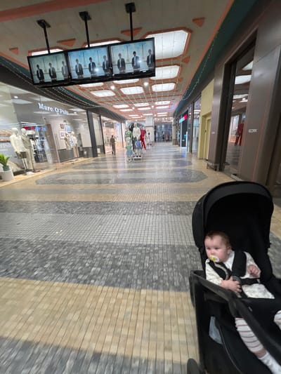 This media file shows a baby in a stroller in a well-lit shopping mall. The baby is wearing a black and white outfit and has a pacifier in its mouth. The stroller is black. The mall appears to have a tiled floor and a long corridor with shops on either side. There are several televisions displaying advertisements featuring men in suits. One shop sign visible says "Marc O'Polo". The lighting suggests it is daytime. The overall scene appears to be a typical shopping environment. The text "BORK" is visible on a sign. Other visible text includes "BORK", "Marc O'Polo", and Russian text that appears to be advertising slogans for stores.