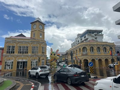 The image captures a street scene in Phuket City Municipality, Thailand, characterized by its Sino-Portuguese architecture. In the foreground, a wet road surface suggests recent rainfall, with cars navigating a roundabout marked by pedestrian crossings. Several vehicles are visible, including a white SUV and a dark sedan.

Dominating the mid-ground is a large inflatable yellow rabbit figure holding a rainbow flag, a prominent display likely for an event. Behind the rabbit, the street stretches into the distance, lined with yellow buildings featuring arcaded ground floors and ornate facades, typical of the region's colonial heritage. A clock tower, also painted yellow, stands tall on the left side of the image, with two clock faces visible.

On the right, a modern white building with a glass facade rises above the older structures. Rainbow flags are also seen hanging from lampposts along the street, further emphasizing a festive or celebratory atmosphere, possibly related to LGBTQ+ pride events. The sky is partly cloudy, with a mix of blue sky and white clouds. People are sparsely visible, some walking and others presumably inside buildings or vehicles.