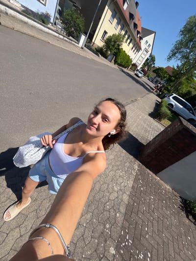 A woman is walking on a sidewalk in a city. She is holding a white bag with a floral pattern and wearing a white tank top and blue shorts. She has a bracelet on her left wrist and is wearing white earbuds. In the background, there are houses, cars, and trees. The image was taken in the city of Saarbrücken, Germany. The sun is shining and the sky is blue. The sidewalk is paved with bricks. The street is asphalt.