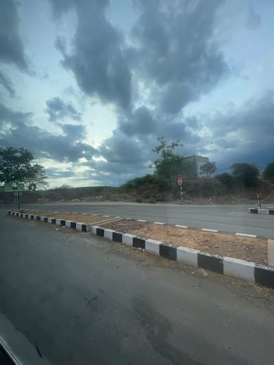 This media file captures a roadside scene near Sugalimetta, India, under a dramatic sky. The image is oriented vertically, with the road and embankment dominating the right side and a cloudy sky filling the left.
The road appears to be paved asphalt, with a rougher, dirt shoulder to its left. A distinct curb runs along the edge of the road, marked with alternating black and white stripes, suggesting a potentially hazardous or visually important area. The embankment beside the road is covered in dry vegetation and sparse trees.
In the middle ground, a white, somewhat weathered building is visible on the hillside, partially obscured by trees. A red octagonal stop sign is planted near the road, indicating a point of control. The sky is filled with dense, dark grey clouds, hinting at impending rain or the aftermath of a storm. The light filtering through suggests it is likely late afternoon or early evening.
The overall atmosphere is one of natural ruggedness and a sense of isolation. No people are visible, and the focus is on the landscape and infrastructure. The stark contrast between the dark clouds and the light filtering through adds a touch of drama to the scene. The presence of the stop sign and the road imply human activity and transportation, but the natural elements, particularly the weather, are a strong visual presence.