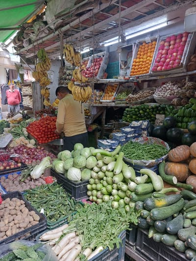 A bustling outdoor market in Manali, India, displays an abundant variety of fresh produce. The scene is dominated by colorful fruits and vegetables arranged in crates and baskets under a green awning. In the foreground, piles of potatoes, okra, and daikon radishes are neatly stacked. Cabbages, gourds, and zucchini fill several crates, creating a vibrant green display. Behind these, a large mound of bright red tomatoes is a central focal point. Bunches of bananas hang from the ceiling, interspersed with other fruits like apples and oranges displayed in boxes. A man in a yellow shirt is visible in the middle ground, appearing to be attending to the produce. Another man in a red sweater stands to the left, observing the market. The environment suggests a lively market day, with the air likely filled with the sounds and smells of commerce.