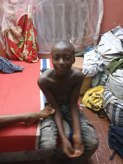 The image captures a young, dark-skinned person, likely a boy, sitting indoors and looking directly at the camera with a neutral expression. He is shirtless and wears dark trousers, seated on a red mattress or mat placed on a reddish-brown tiled floor. His hands are clasped in front of him.
To the boy's left, a white mosquito net is draped over what appears to be a sleeping area. A red fabric with large white and green floral patterns is neatly folded on the mattress, below the mosquito net. Another piece of fabric, dark blue with white stripes, lies on the mattress in the lower left foreground.
Behind the boy, various clothes and personal items are piled up on the floor or an unseen surface. These include light-colored garments, a green fabric, a black and white striped bag or textile, and other dark blue clothing. A yellow backpack with brown straps is also visible among these belongings. Part of a bare foot, possibly belonging to another person, is visible on the right side of the frame, resting near the edge of the mattress.
The setting suggests a simple indoor living space or bedroom in Idah, Nigeria. The lighting is moderate, indicating either daytime or an illuminated evening. No visible text is present in the image.