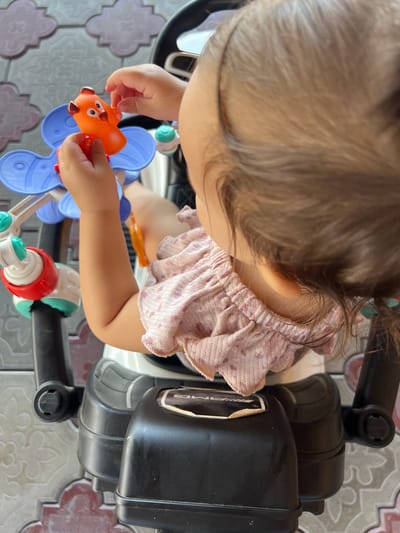 A close-up shot shows a young child's hands interacting with a bright orange toy animal, possibly a dog, with large ears and polka dot markings. The toy is attached to a blue plastic structure with petal-like extensions. The child is seated in a black ride-on toy, possibly a stroller or tricycle, with visible red and green components of another toy on the left. The child is wearing a pink, ruffled top. The background features a patterned floor with light grey and pink tiles. The image is taken from a high angle, focusing on the child's activity and the toys. The setting appears to be outdoors or in a well-lit indoor space with a patterned tiled floor. The location is identified as Qoyankus, Kazakhstan, though this specific detail cannot be visually confirmed from the provided image.