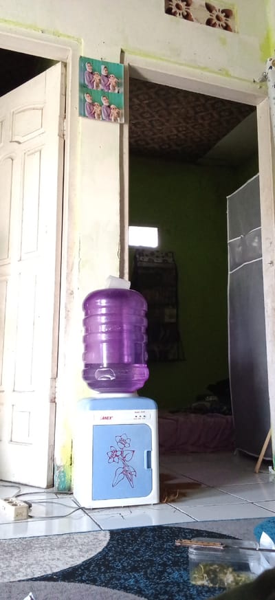 The media file captures an indoor domestic scene, likely a hallway or entryway, opening into another room. The focal point is a purple plastic water dispenser bottle, approximately half full, resting on a compact, light blue and white water cooler or mini-fridge. A small white paper cup sits inverted on top of the water bottle.
To the left, a white wooden door is partially ajar, revealing a glimpse of the bright outdoors or another brightly lit area. On the white wall above the water dispenser, a laminated picture is affixed, displaying two identical rows of a woman wearing a head covering, holding two monkey dolls.
Through the open doorway behind the water cooler, an interior room with light green painted walls is visible. Bright sunlight streams in through a small, high rectangular window on the far wall of this room. Below the window, a dark-colored shelf unit or rack can be seen. Further into the room, a bed with a pink sheet is partially visible, and to the right, a tall, gray fabric wardrobe or storage unit stands. The ceiling of this inner room appears to have a patterned, possibly textured, finish.
The floor in the foreground is tiled with light-colored squares, showing some cracks. A patterned rug with dark blue, gray, and white abstract shapes lies on the floor. Near the bottom left corner, a white power outlet strip is connected to cords. On the rug, a clear plastic container holds some green, leafy material, and a small wooden implement rests on top of it.
The setting appears to be daytime due to the bright natural light. Visible text on the water cooler includes the brand name "SANEX" and "Model: DS-XX" (the last two digits are indistinct). No human figures are present, suggesting a quiet moment in a residential environment.