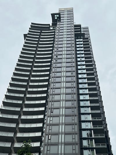 A striking image captures a modern, tall building from a low-angle perspective, rotated so the structure extends diagonally across the frame against an overcast sky. This building, situated in Coquitlam, Canada, showcases a distinct architectural design.

The upper portion of the building features a more traditional façade of dark-colored panels and rectangular windows arranged in a uniform grid. Below this, a more dynamic section emerges, characterized by a series of stepped or layered balconies or architectural projections. These elements extend outwards, creating a textured, multi-dimensional effect. Each projection is composed of dark panels and windows, angled slightly, enhancing the building's visual depth. The cropped images further detail these repetitive architectural patterns, highlighting the sleek lines and dark materials.

The building's exterior is predominantly dark, suggesting materials like glass, metal, or dark stone, which reflect the soft, diffused light of the cloudy day. The overall color palette is largely monochromatic, dominated by shades of grey and black. The sky is uniformly light grey and overcast, indicating a cloudy daytime. A small cluster of green foliage, likely from a tree or bush, is visible at the bottom right of the image, providing a natural element against the urban structure. There is no visible text.