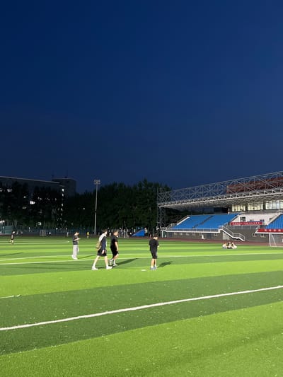 The image depicts an outdoor sports field, likely a soccer or track and field stadium, at dusk. The sky is a deep, dark blue, indicating the transition from day to night. The field itself is artificial turf, divided into alternating shades of green with white lines marking boundaries and lanes. In the background, blue stadium seating is visible, along with a metal support structure for lights or a roof. Trees line the edge of the field, and a building with illuminated windows can be seen further in the distance, suggesting an urban or suburban setting.

Several people are present on the field, seemingly engaged in physical activity. One person in a black t-shirt and shorts stands on the field, casting a shadow. Further away, a group of individuals are seen in various poses, some appearing to be stretching or resting. The overall atmosphere suggests a training session or a casual gathering after sporting events. The lighting is a mix of natural twilight and artificial stadium lights which are not clearly visible but illuminate the field.

A visible sign on the stadium seating displays Chinese characters, which translate to "Daxing District 2023 Sports Meeting" and "Tianjin University of Science and Technology". The presence of this text clearly indicates the location and the context of the event as a sports meeting.
