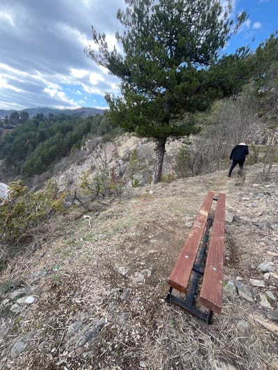 The image displays a rugged hillside view in Srednogortsi, Bulgaria, under a sky with both blue patches and elongated grey clouds. In the immediate foreground, a wooden bench with two reddish-brown slats and black metal supports is situated on uneven ground composed of dry grass, dirt, and scattered rocks. The left slat of the bench shows some surface markings or carvings. A lone individual, seen from behind, is walking uphill and to the right on a discernible dirt path. This person is dressed in a dark jacket with a visible light-colored lining around the hood/collar, dark trousers, and dark footwear. To the left of the path, a prominent pine tree with dense green needles stands. The midground features a steep, rocky slope descending to the left, at the base of which a segment of a paved road is visible. Beyond this, a vast coniferous forest covers multiple rolling hills, with distant mountains and faint architectural structures appearing on the horizon. The surrounding environment includes numerous bare deciduous trees and shrubs, suggesting a late autumn, winter, or early spring period.