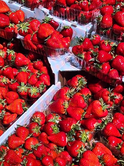 The image shows a close-up view of numerous plastic containers filled with ripe strawberries, displayed in what appears to be a market or roadside stand. The strawberries are a vibrant red, with some showing green leaves and stems. The containers are stacked and arranged, suggesting a retail setting. The lighting indicates it is daytime, and the overall impression is one of fresh produce being sold. There are no people visible in the frame, and no specific landmarks that would pinpoint a location within Voluntari, Romania. The image focuses solely on the strawberries and their packaging.