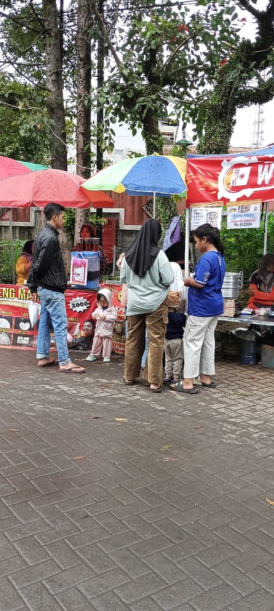 The scene is a street food market in Banjaran, Indonesia. Several people are standing around a food stall. There are two umbrellas, one red and one multicolored. The stall is selling food and drinks, including "tahu baso" for Rp. 2000. One man is wearing a black jacket and blue jeans, another woman is wearing a green hijab, and another woman is wearing a blue shirt. A young girl with a pink shirt is standing next to the stall with a man. In the background, a woman is sitting next to another food stall wearing a red shirt. Behind the stall is a building and some trees. The ground is paved with gray bricks.
