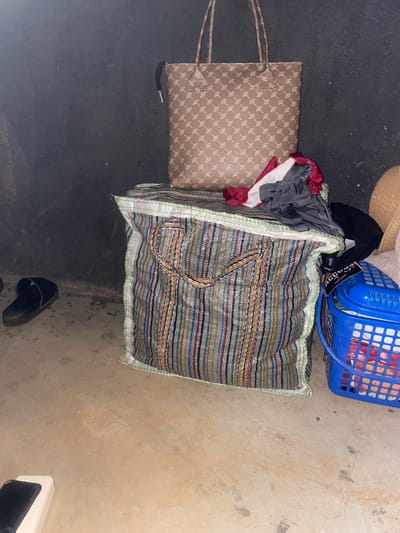 An indoor scene depicts a cluttered floor area, possibly in a storage space or room in Unknown, Nigeria. A large, colorful woven bag with a striped pattern is prominent in the foreground. To its left, a black sandal rests on the concrete floor. Behind the woven bag, a smaller, tan-colored tote bag with a repeating geometric pattern is placed on top of a pile of clothes, which include red, gray, and white fabric. To the right of the woven bag, a blue plastic basket filled with items, some appearing red, is visible. A light-colored hat is partially visible to the right of the basket. The background consists of a dark, textured wall. The overall impression is one of disorganized storage.