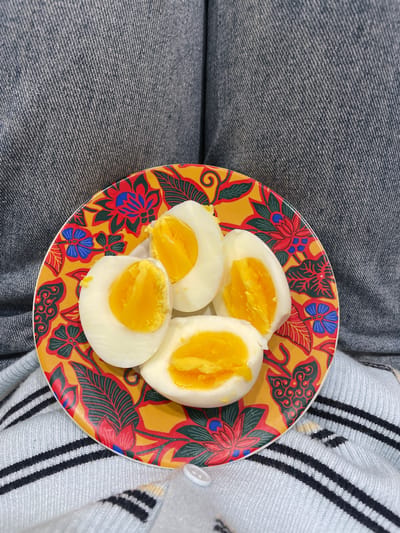 A plate of four hard-boiled eggs, halved and arranged on a decorative yellow plate with a floral pattern, is presented on a surface covered by blue denim and a light blue and white striped knitted garment. The eggs have firm white albumen and vibrant yellow yolks, some of which are slightly moist. The plate's design features red, blue, and green floral motifs and black swirling lines. The background consists of textured blue denim, possibly part of a pair of jeans, and the striped fabric of the garment. The scene is indoors, with no discernible city landmarks or specific urban features of Bekasi, Indonesia. The lighting is diffused, suggesting an indoor environment.