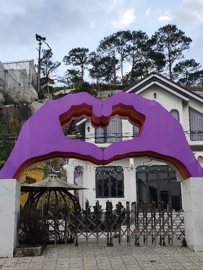 The image captures an exterior view in Thành phố Đà Lạt, Vietnam, centered on a large, purple archway designed as two hands forming a heart. The archway features interior concentric bands of orange, yellow, and red. It is situated on a paved surface composed of light-colored interlocking tiles.

Behind the archway, a two-story white building with a brown tile roof and prominent dark-framed windows is visible. Some windows display grey curtains, and multiple exterior wall sconces are affixed to the facade. A fully extended, accordion-style stainless steel gate blocks access to the building's entrance. Overhead string lights are strung across the building's exterior, positioned above the gate. To the left of the gate, a weathered, round, umbrella-like structure, possibly a gazebo roof, is partially visible, alongside a desiccated plant in a dark planter.

The background features a steep hillside densely covered with numerous mature pine trees. A white-framed greenhouse structure is observable on the upper left slope. The sky is largely overcast. No human figures are present within the frame.
