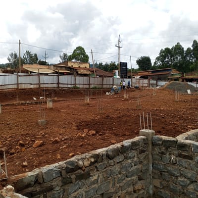 The image captures an active construction site in Borabu Chitago ward, Kenya, under a cloudy, overcast sky during the daytime. In the immediate foreground, a robust stone foundation wall, constructed from dark, irregularly shaped stones, is partially built. Beyond this, the ground is a expanse of reddish-brown soil, indicative of recent excavation, dotted with rocks and partially completed concrete pillars with rebar extending upwards, marking the future structure's footprint.
In the mid-ground, construction activity is underway with a blue concrete mixer visible. Several workers are present; three men are standing near the mixer, with one appearing to operate it, while another worker stands further to the right. Behind the construction area, a corrugated metal fence separates the site from the adjacent properties.
The background reveals a local commercial or residential area with various buildings. These structures generally feature corrugated iron roofs and simple facades. Visible text on a building to the left reads "SEASONS SPECIAL QUALITY HOSTEL." Further to the right, another building displays a sign for "ALLE AGROVET" and a smaller "m-pesa" sign beside it. Wooden utility poles with power lines stretch across the frame, connecting to the nearby buildings. Lush green trees are scattered throughout the background, adding to the semi-urban landscape. The scene conveys a sense of ongoing development and community life.
