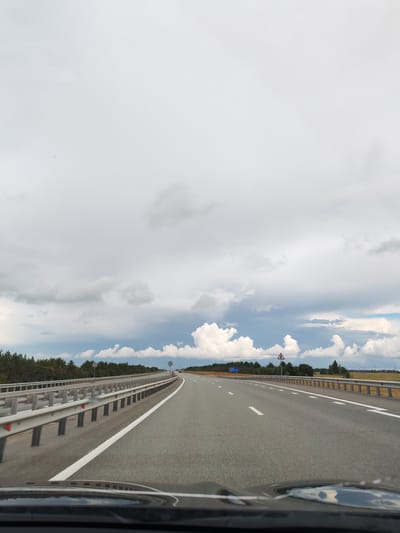 The image shows a view from a vehicle traveling on a highway. The road is paved and has white lane markings. On the left side of the road, there is a metal guardrail. On the right side of the road, there is more guardrail and then a field. In the distance, there are trees and a cloudy sky. There are no people visible in the image. The location is stated to be the city of Kzyl-Tan, Russia, but this cannot be confirmed from the image itself.