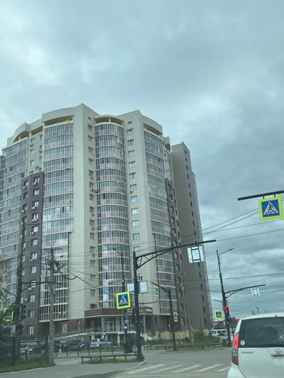 The image shows a modern, multi-story residential building with a beige and dark brown facade and numerous windows. The building is situated in the city of Yakutsk, Russia. In the foreground, there is a street with a pedestrian crossing, traffic lights, and overhead electrical wires. A white car is visible in the lower right corner of the frame. The sky is overcast with grey clouds.
