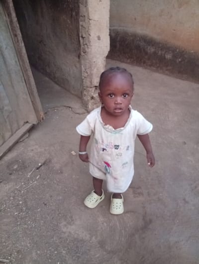 A young child stands on a gray concrete surface in front of a building with a rough exterior. The child wears a white, short-sleeved garment and white shoes. A bracelet adorns their left wrist, and they hold a thin object in their right hand. The background suggests an outdoor setting, possibly near a residence or building structure with rustic elements. The child gazes forward with a slightly curious expression.