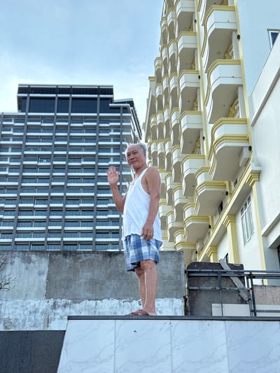 A man in a white tank top and blue and white checkered shorts stands on a tiled surface, with his right arm extended and hand open, as if waving. He is facing the camera and appears to be smiling. Behind him, a modern high-rise building with many windows dominates the background. To the left of the man, a lighter-colored building with ornate architectural details, possibly a hotel or resort, is visible. The scene appears to be outdoors, with a cloudy sky. The overall impression is a casual, relaxed moment captured during the daytime.
