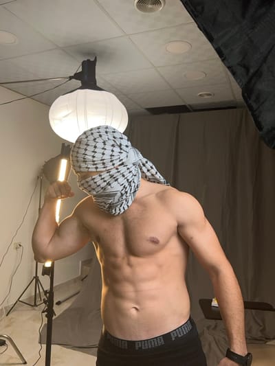 A shirtless man with a keffiyeh wrapped around his head poses in a studio setting. He has a muscular physique, and is wearing black underwear with a Puma logo. The scene includes photography equipment, with a softbox and an LED light panel visible. The walls and ceiling of the studio are visible in the background.