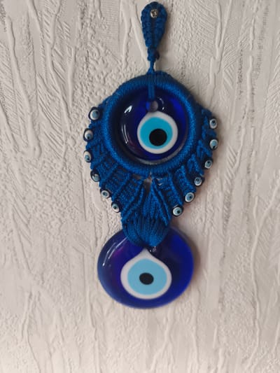 This is a close-up, vertical, eye-level shot of a blue evil eye wall hanging. The evil eye is a decorative object, often used as a talisman, believed to ward off evil spirits and protect from misfortune. It is made of glass and yarn, with a beaded decorative element. The evil eye is suspended from a small loop of blue yarn that is attached to a screw protruding from a textured white wall. The wall appears to have a raised, linear pattern, possibly wallpaper or a textured paint finish. The lighting suggests it is daytime, with soft light illuminating the object and its surroundings. The image is centered on the evil eye, with the textured wall filling the background. There is no visible text in the image. The setting appears to be indoors, possibly a home in Eitorf, Germany. The overall mood is neutral, focusing on the details of the object and its presentation.