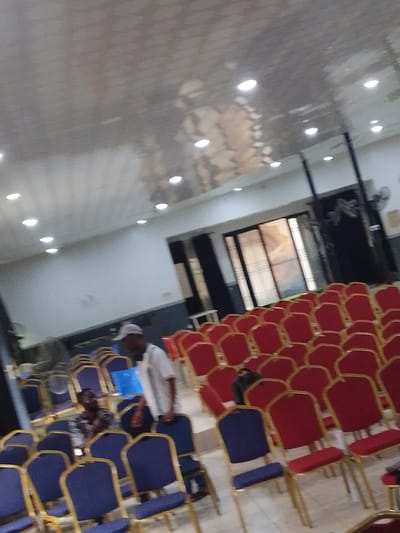 This image depicts the interior of a hall, possibly a church or event space, in Osogbo, Nigeria. Rows of chairs, predominantly red with gold frames, fill the space. Some blue chairs are also visible. A man wearing a white t-shirt and a grey baseball cap stands among the chairs, facing away from the camera. Another person is seated in a blue chair, also with their back to the camera. The ceiling is a metallic, reflective surface with multiple recessed lights. A large sliding door or window is visible in the background, with a dark framed structure. There are also industrial fans mounted on the wall. The lighting appears to be from the ceiling lights and possibly ambient light from the background. The floor is tiled.