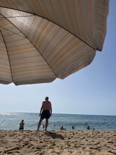 An outdoor, eye-level, medium shot captures a beach scene in Badalona, Spain, under a clear blue sky. In the foreground, a large, striped umbrella casts a shadow over the sandy beach, its fabric displaying horizontal bands of pink, orange, yellow, and gray. The umbrella is angled to fill the top third of the frame, with its ribbed structure visible. In the mid-ground, a man wearing dark swim trunks stands on the sand, facing the ocean, his back to the camera. To his left, a woman in a blue bikini sits on the sand, also facing the water. The ocean stretches across the background, shimmering with sunlight, and several people are visible swimming and wading in the water. The sand is a light brown color, with visible texture and some debris. The overall impression is a sunny day at the beach.