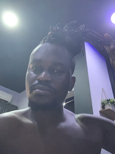 An adult dark-skinned male is centered in a dimly lit indoor setting, depicted from the chest upwards. He has a short beard and mustache, with dark dreadlocks styled in a bun on his head. His left hand is visible near the top right, appearing to adjust or touch his dreadlocks. He is shirtless and looks directly at the camera with a neutral expression. The background features a dark ceiling with two circular lights: a bright white light on the upper left and a dimmer purplish light on the upper right. A white wall-mounted air conditioning unit is partially visible on the left. On the right, a wall exhibits a purple ambient light, below which a wooden shelf supports a decorative plant with green foliage housed in a geometric, possibly metallic, frame. The specific location within Jibowu, Nigeria, is not discernible from the image content.