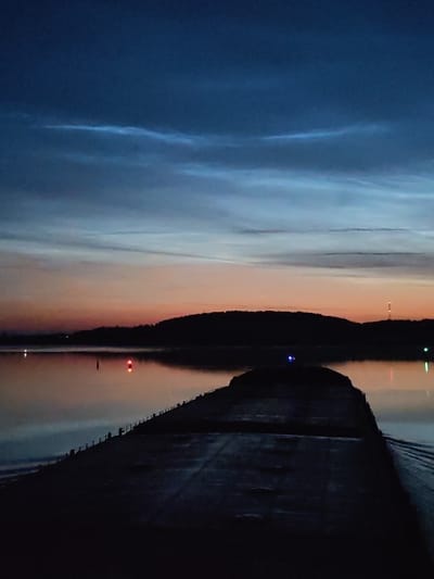 This image depicts a twilight scene over the Zvoz River in Zvoz, Russia. The sky is a gradient of deep blue at the top transitioning to a soft orange and pink near the horizon, with wispy, luminous clouds, possibly noctilucent clouds, visible in the upper portion. A dark, silhouetted landmass with a tree line forms the background. In the foreground, a dark, concrete structure, possibly a pier or jetty, extends into the water. The water reflects the colors of the sky and shows several lights: a red navigational buoy, a faint white light, and a blue light near the silhouetted landmass. The overall impression is one of a serene, late evening or early morning landscape.