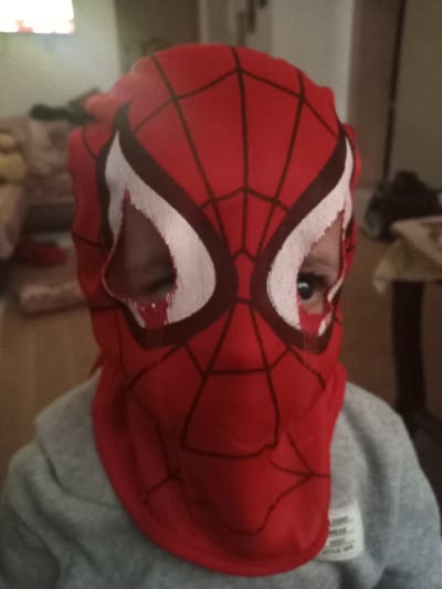 An individual, likely a child, is depicted in a medium close-up, facing directly towards the camera. The person is wearing a red fabric mask designed to resemble Spider-Man, featuring black web patterns and white eye sections outlined in dark red. The mask covers the entirety of the head and neck, with only the subject's eyes visible through irregular openings. Below the mask, a grey hooded sweatshirt is partially visible, with a white textile label displaying black text on the right chest area. The background is an out-of-focus indoor environment, presenting a light-colored sofa or armchair to the left and indistinct furniture or objects to the right. A soft, diffused light source is present in the upper left background. The scene is located in a residential or interior space within Dar Bouazza, Morocco.