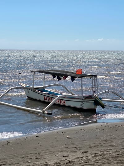 A white outrigger boat named "KETUT ONTOST" is partially submerged in the shallow, choppy, sunlit ocean waters near the sandy shore of Buleleng, Indonesia. The boat has a dark canopy supported by white poles and a red flag is visible on the canopy. A green covering is draped over the stern of the boat. The ocean water is a deep blue with shimmering highlights from the sun. The beach is composed of dark sand with scattered debris. The sky above is a clear, bright blue.