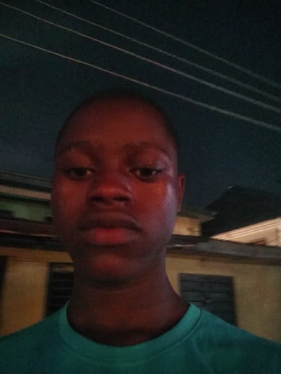 A close-up, low-angle, front-facing selfie of a dark-skinned young person wearing a teal t-shirt is taken outdoors at night. The background consists of out-of-focus, dimly lit buildings and electrical wires against a dark sky. The subject's face is in focus, with their eyes, nose, and mouth clearly visible. The lighting highlights the sheen on their skin, suggesting sweat or oil. The image appears to be taken in Shangisha, Nigeria, based on the contextual information provided.