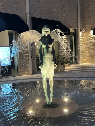 This media file depicts a bronze statue of a person with wings, positioned within a modern architectural setting. The statue is partially submerged in water, which is flowing around it, creating a dynamic visual effect. The water appears to be part of a fountain, with streams cascading and splashing.
The setting features a building with a textured, light-colored stone facade and vertical architectural elements. There is a sign visible with text that partially reads "TON REAL ESTATE". The lighting suggests it is either dusk or nighttime, as artificial lights are illuminated, casting a warm glow on the stone and highlighting the water. One light source appears to be embedded in a circular stone base where the statue's feet are positioned.
The statue itself is a striking feature, with a weathered bronze patina. The figure has a serene or perhaps contemplative expression. The wings are rendered in a way that suggests movement and the flow of water.
The overall impression is one of a public art installation in a contemporary urban or commercial environment. The combination of the sculpture, the water feature, and the architecture creates an artistic and serene atmosphere. There are no clear indicators of specific weather conditions, but the focus is on the interplay of light, water, and sculpture.