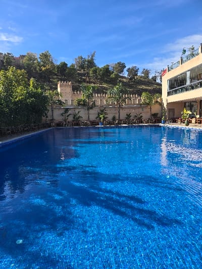 The image presents an outdoor swimming pool area in Taza, Morocco, under a clear blue sky. A large, rectangular swimming pool with visible blue mosaic tiling fills the lower portion of the frame, its surface reflecting sunlight and displaying ripples. Along the left edge of the pool, several dark-colored chairs are arranged. In the midground, a light-colored, crenellated fortified wall, featuring at least one square tower, extends horizontally, separating the pool area from a densely vegetated hillside covered with green trees. Potted plants and outdoor seating arrangements (tables and chairs) are situated along the fortified wall and near the pool. To the right, a multi-story, modern building with beige facades, extensive windows, and balconies is visible. A red flag with a green pentagram, identified as the Moroccan flag, flies from the upper section of this building. No individuals are present in the scene.