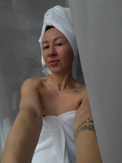 A woman with a white towel wrapped around her head and body takes a selfie in what appears to be a bathroom. Her left arm is extended, holding the camera. She has a tattoo on her right upper arm, which appears to be a floral or vine design. The background is blurred and shows a light-colored wall with some texture, and a sheer curtain partially obscures the view. The lighting is soft and diffused. The location is identified as Novaya Vozhoika, Russia.