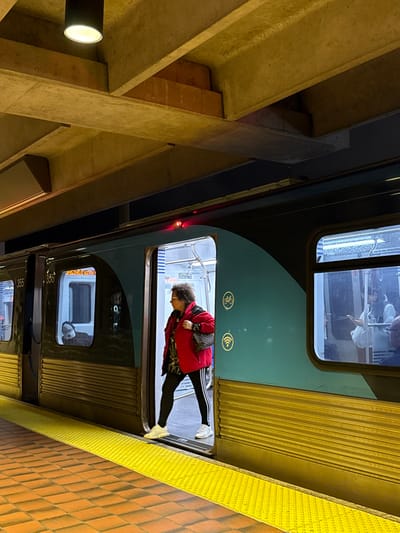 A train car, oriented vertically, shows a woman in a red jacket entering or exiting. Inside, people are reflected in the windows. Outside, a textured yellow platform edge is visible. Adjacent to the train is a light-colored concrete structure with visible beams and a light fixture. The image is taken at an angle.