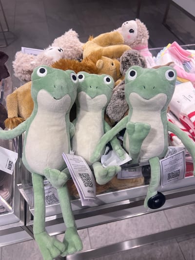 A shelf displays a variety of stuffed animals and infant apparel. Three green frog plush toys dominate the upper right, each bearing a white tag with black print and a QR code. To the left of the frogs are several smaller, indistinct stuffed animals in beige, brown and cream colors. At the top of the frame are stacked pink and white infant socks. The scene appears to be inside of a retail store. There is no evidence to support a connection to the city of San Pedro Cholula, Mexico.