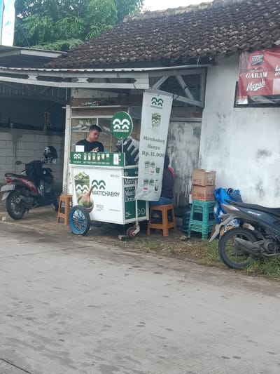 A small "MATTCHABRY" matcha drink stand is set up roadside, with a worker behind it. The stand is white, branded with the "MATTCHABRY" logo, and features a menu. A banner displays "Matchaboy Hanya Rp. 12.000" (Matchaboy Only Rp. 12,000). A blue decorative wheel is affixed to the side. A customer sits on a stool. Two motorbikes are parked beside the stand. A house with a tiled roof provides a backdrop. A sign in red reads "Gudang Garam Merah."