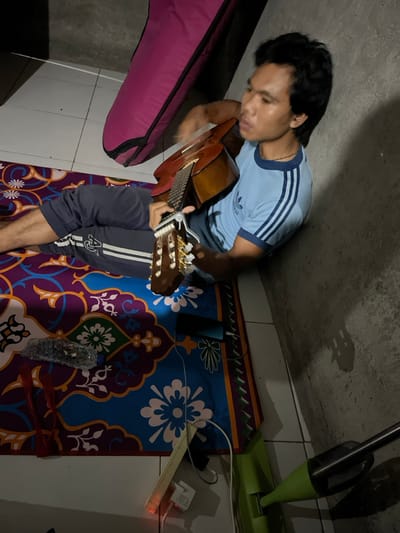 An adult male is observed sitting on a multi-colored patterned mat on a tiled floor in Desa Batu Layar, Indonesia. He is leaning against an unpainted concrete wall. The man wears a light blue t-shirt with dark blue trim and three stripes on the sleeves, and dark grey sweatpants with a white logo and stripe on the left leg. He is barefoot. He holds a light brown acoustic guitar, positioning his left hand on the fretboard where a silver capo is attached, and his right hand near the soundhole, indicating he is playing. His head is turned slightly to the right, and his lips are slightly pursed. To his left, a pink zipped guitar case leans against the wall. On the mat, a black smartphone is visible, connected to a white charging cable. A white power strip, illuminating with an orange light, rests partially on the mat and tiled floor, connected to other white cables and chargers. A clear plastic bottle containing miscellaneous debris, along with some red string, lies on the mat. In the lower right corner, a portion of a green and black cleaning tool is visible. The scene is illuminated by an unseen light source, casting shadows on the concrete wall.