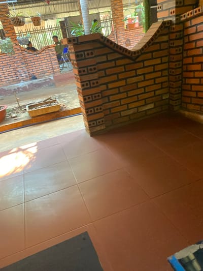 The image, taken in Đak Đoa, Vietnam, captures a transitional space between an indoor area and an outdoor patio. The immediate foreground features a reddish-brown tiled floor, with a dark rectangular mat in the lower-left corner and a blue object partially visible in the lower-right. A strong sunlit reflection illuminates the left side of the tiled floor. Dominating the right side of the frame is a brick partition wall constructed from red-brown bricks with dark mortar, featuring a distinctive M-shaped crest along its upper edge.
Beyond this partition, through an open passage, an outdoor area is visible. The ground is a mix of dirt and concrete, with a blue pipe running horizontally along its edge. A brown potted plant is on the far left, and a rectangular wooden planter box filled with dry leaves or debris rests near the center-left. Further back, a decorative brick wall, also of red-brown bricks with a wavy top pattern, serves as a barrier. Overhead, a corrugated metal roof structure supports numerous hanging plants in terracotta and dark baskets.
Two individuals are present in the outdoor area. One person, wearing a light green long-sleeved shirt and dark pants, stands near a white folding chair, facing left and partially obscured by the foreground brick wall. Another person is partially visible behind the wavy brick wall, to the left of the first individual. A metal fence and dense green foliage are visible in the background beyond the roofline.