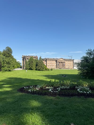 The image depicts a view of the Orangerie in Kassel, Germany, under a clear blue sky. The grand Orangerie building, characterized by its classical architecture with a colonnaded facade, stands prominently in the mid-ground. To the left of the Orangerie, another building, possibly part of the same complex or a separate historical structure, is visible, featuring a more angular design.
The foreground is dominated by a lush green lawn, meticulously maintained. A flowerbed with white and purple flowers is situated in the lower part of the frame, adding a touch of color to the scene. Several trees, including tall, mature ones, are scattered across the lawn and frame the buildings, providing shade and a natural element to the landscape.
In the mid-ground, a single person is walking across the lawn, dressed in light-colored clothing. Further to the right, a group of people are seated on the grass, appearing to be enjoying a picnic or simply relaxing in the park. In the distance, behind the Orangerie, construction scaffolding and a crane indicate ongoing building work. Benches are visible on the lawn in front of the Orangerie, suggesting this is a public space for recreation and enjoyment. The overall impression is one of a serene and well-kept park surrounding a significant historical landmark.