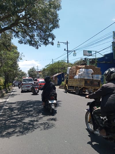 The scene depicts a daytime view of a multi-lane asphalt road in Tagtse Tashidingkha, Bhutan, under a clear blue sky. In the foreground on the right, a person wearing a dark helmet and clothing is partially visible, operating a dark motorcycle. To the immediate left, a second individual, also wearing a dark helmet and clothing, rides a dark scooter with a white rear license plate (partially visible, "52xx"). Dappled shadows from large trees lining the left side of the road are cast across the pavement. Further ahead on the right, a yellow flatbed truck is heavily laden with numerous brown cardboard boxes and several large white sacks, stacked high above the vehicle's cab. In the mid-ground on the left lane, a white ambulance with red "AMBULANCE" text is visible, followed by a red vehicle and other traffic receding into the distance. The right side of the road features overhead utility lines, black streetlights with ornate, cage-like lanterns, and commercial-style buildings partially obscured by foliage.