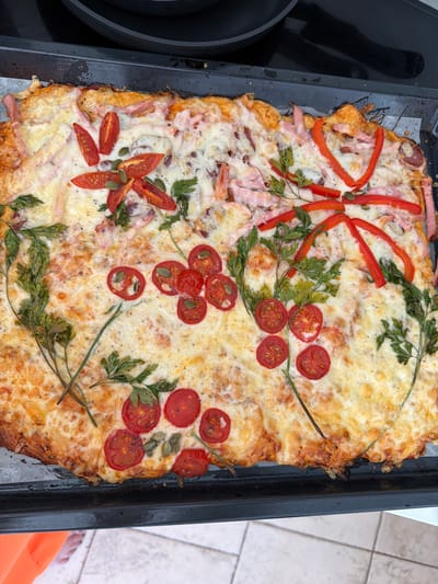 The image shows a close-up, overhead view of a rectangular, thin-crust pizza baked on a black metal baking sheet. The pizza is topped with melted cheese, sliced red bell peppers arranged in a bow-like pattern on the left side, and halved cherry tomatoes. Green parsley sprigs and pumpkin seeds are scattered across the pizza. Some slices of ham are visible beneath the cheese. The pizza has a golden-brown crust, with some darker, slightly charred edges on the right side.
The setting appears to be an outdoor tiled patio, suggested by the tiled flooring visible to the right of the baking sheet. The lighting indicates it is daytime. The scene is a food preparation or serving context. There is no visible text in the image. The focus is entirely on the pizza, which is presented as a savory dish.