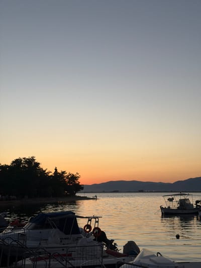 A serene evening scene unfolds at the harbor of Keramoti, Greece, with the sky transitioning from a pale blue to a soft orange hue near the horizon. The tranquil waters of the Aegean Sea gently reflect the warm tones of the sunset. In the foreground, several small boats are moored, their silhouettes distinct against the water. One boat in particular, a white vessel with a blue canopy, is prominent, with life preservers attached to its side. Further out in the water, a larger fishing boat with a canopy is anchored. The shoreline to the left is lined with dark trees, suggesting a coastal vegetation. Silhouettes of distant mountains are visible on the horizon, completing the picturesque landscape. A few individuals are faintly visible on the shore, observing the sunset. The overall atmosphere is one of peacefulness and natural beauty.
